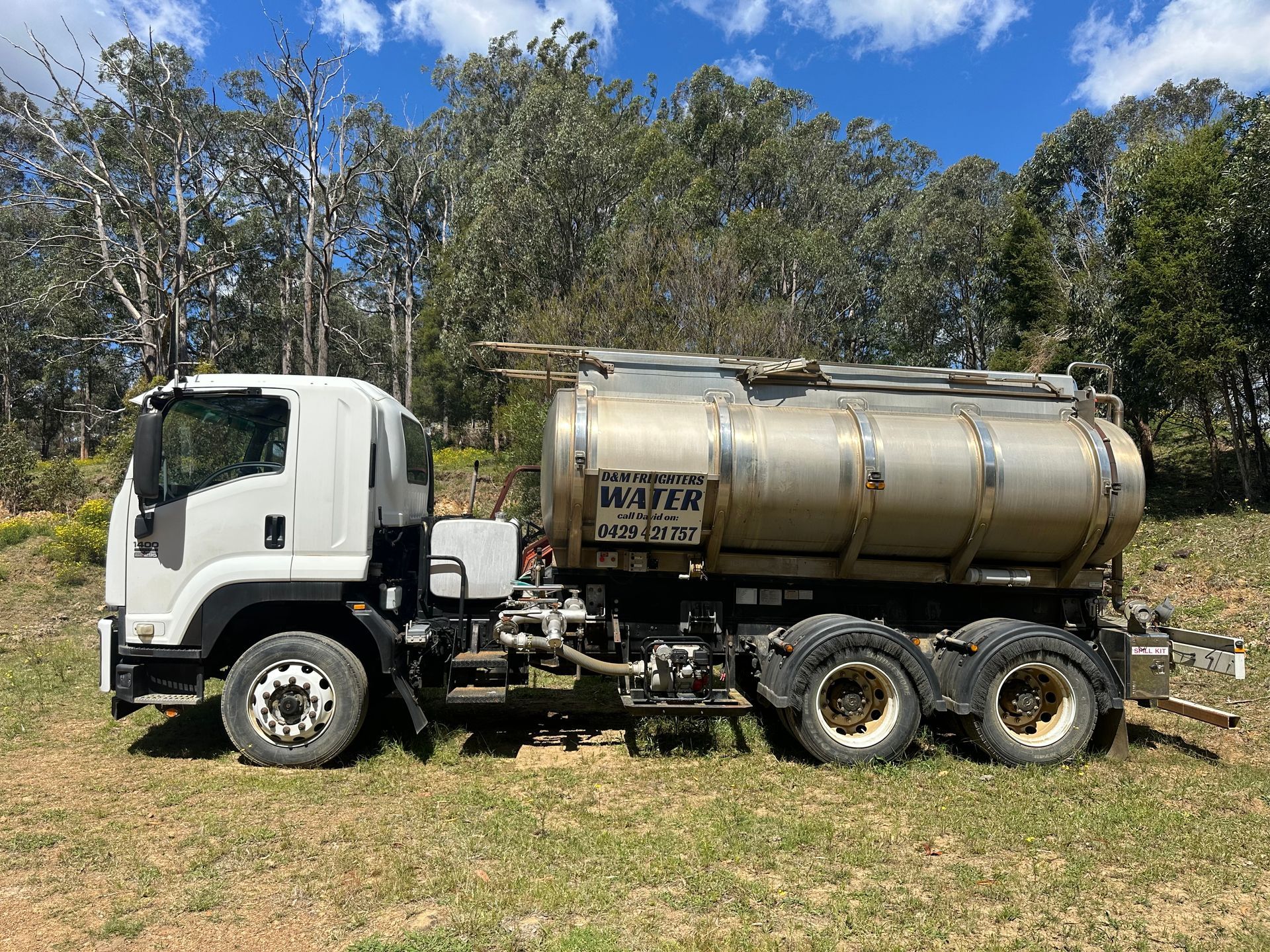 White water truck with a large silver tank parked on dry grass, trees in the background — D & M Freighters Water In Bucca Wauka, NSW