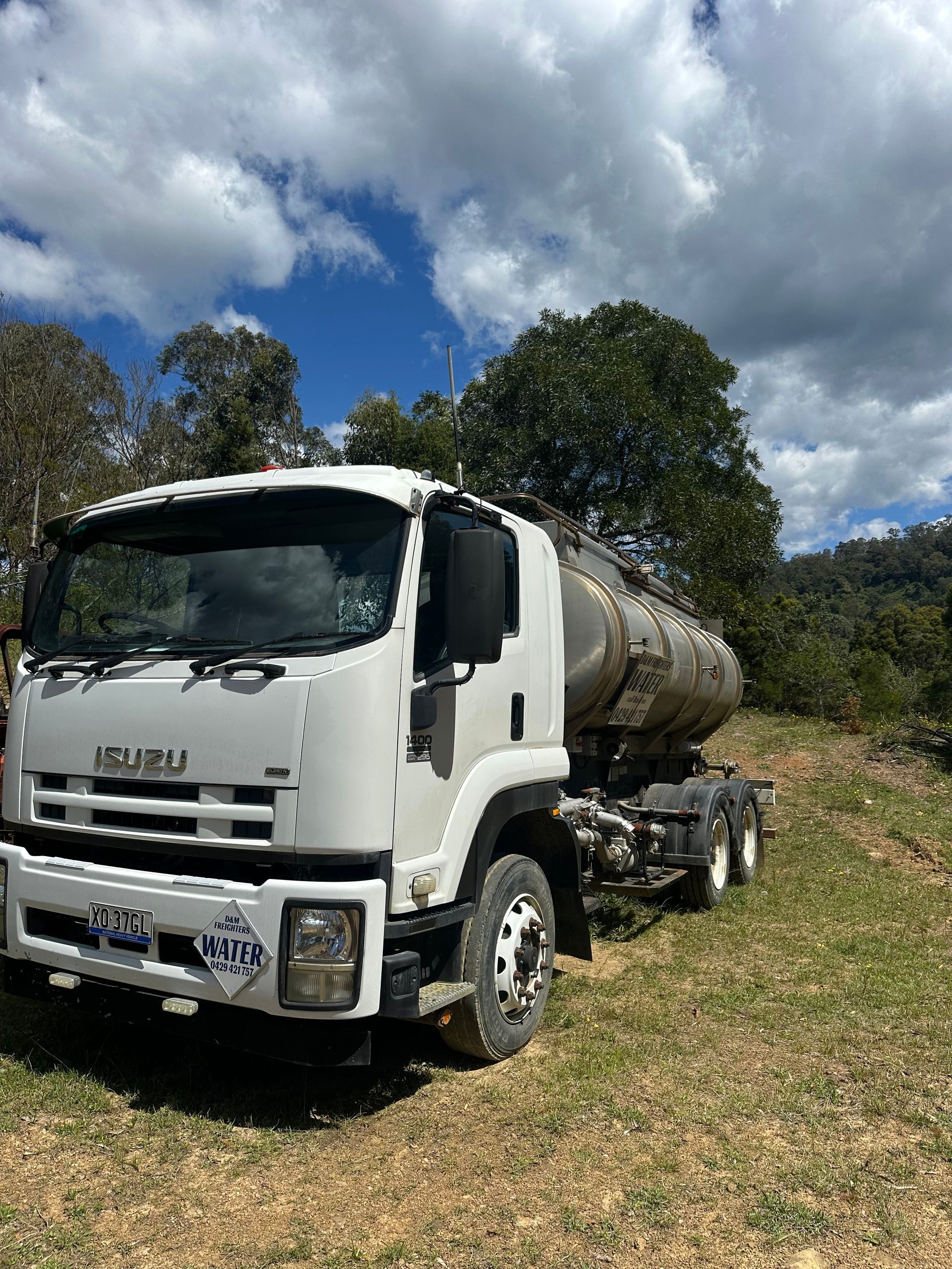 White Isuzu water tanker truck on a grassy dirt road, cloudy blue sky background — D & M Freighters Water In Bucca Wauka, NSW