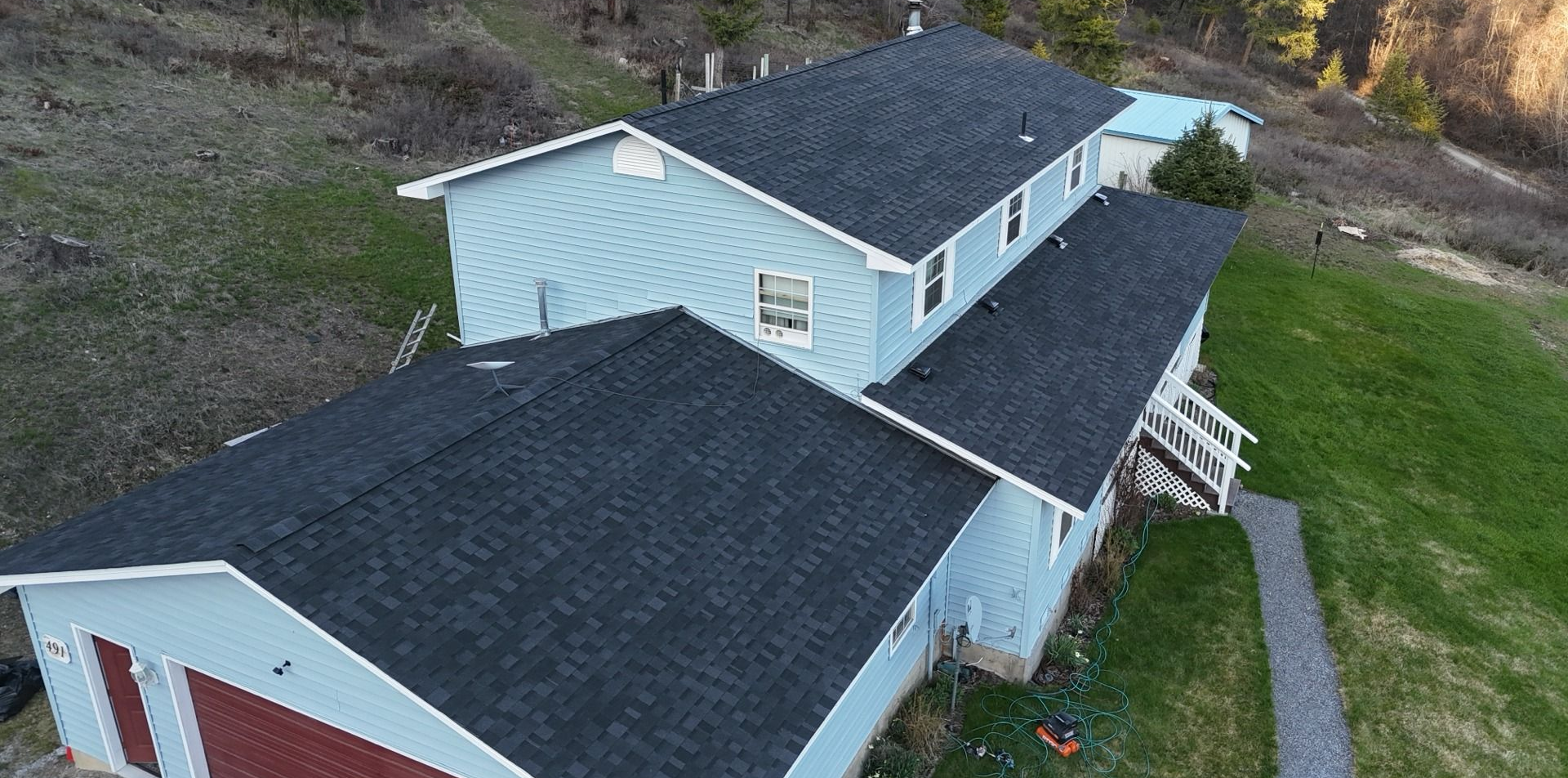 Aerial view of a light blue house with dark roofs beside a grassy yard and gravel path.