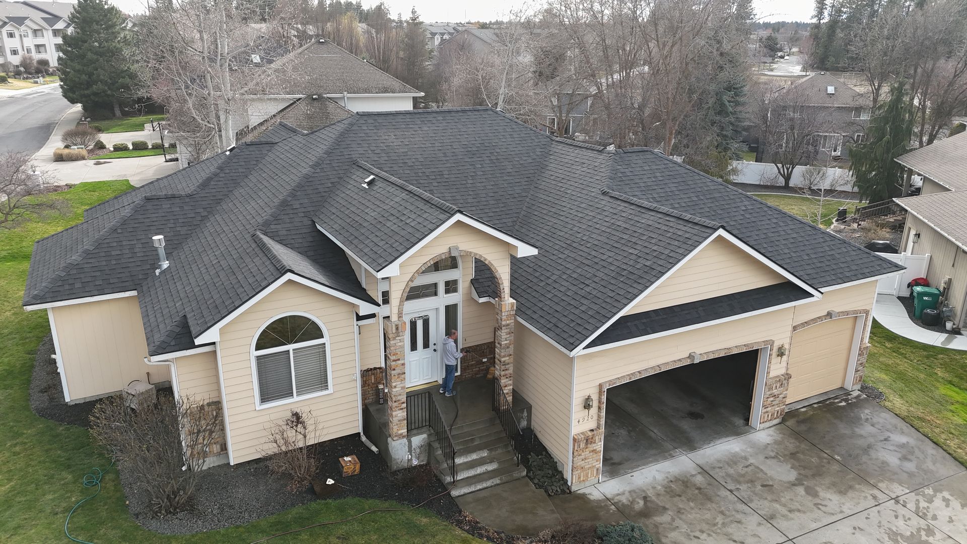 Aerial view of a beige suburban house with a dark roof, front walkway, and attached garage.
