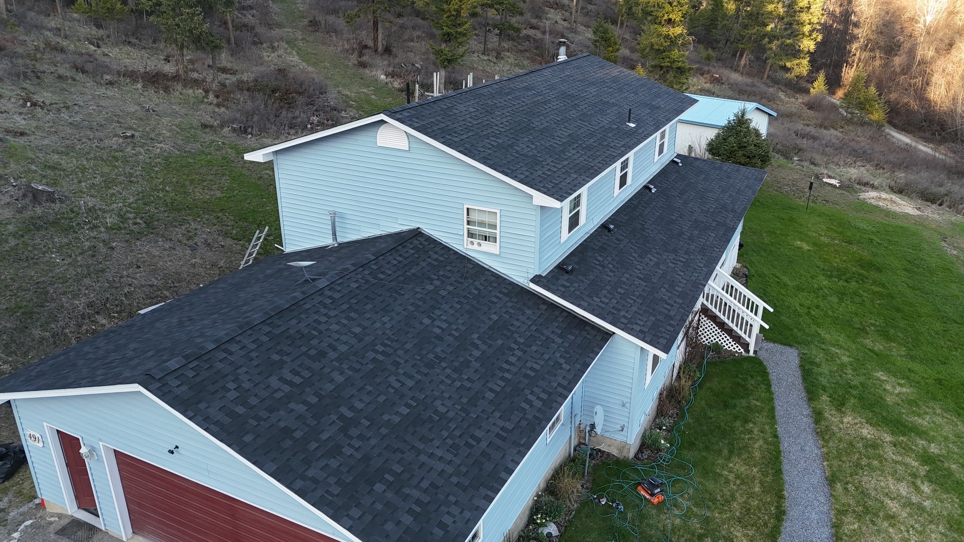 Aerial view of a light blue house with dark roof beside a grassy yard and wooded hillside.
