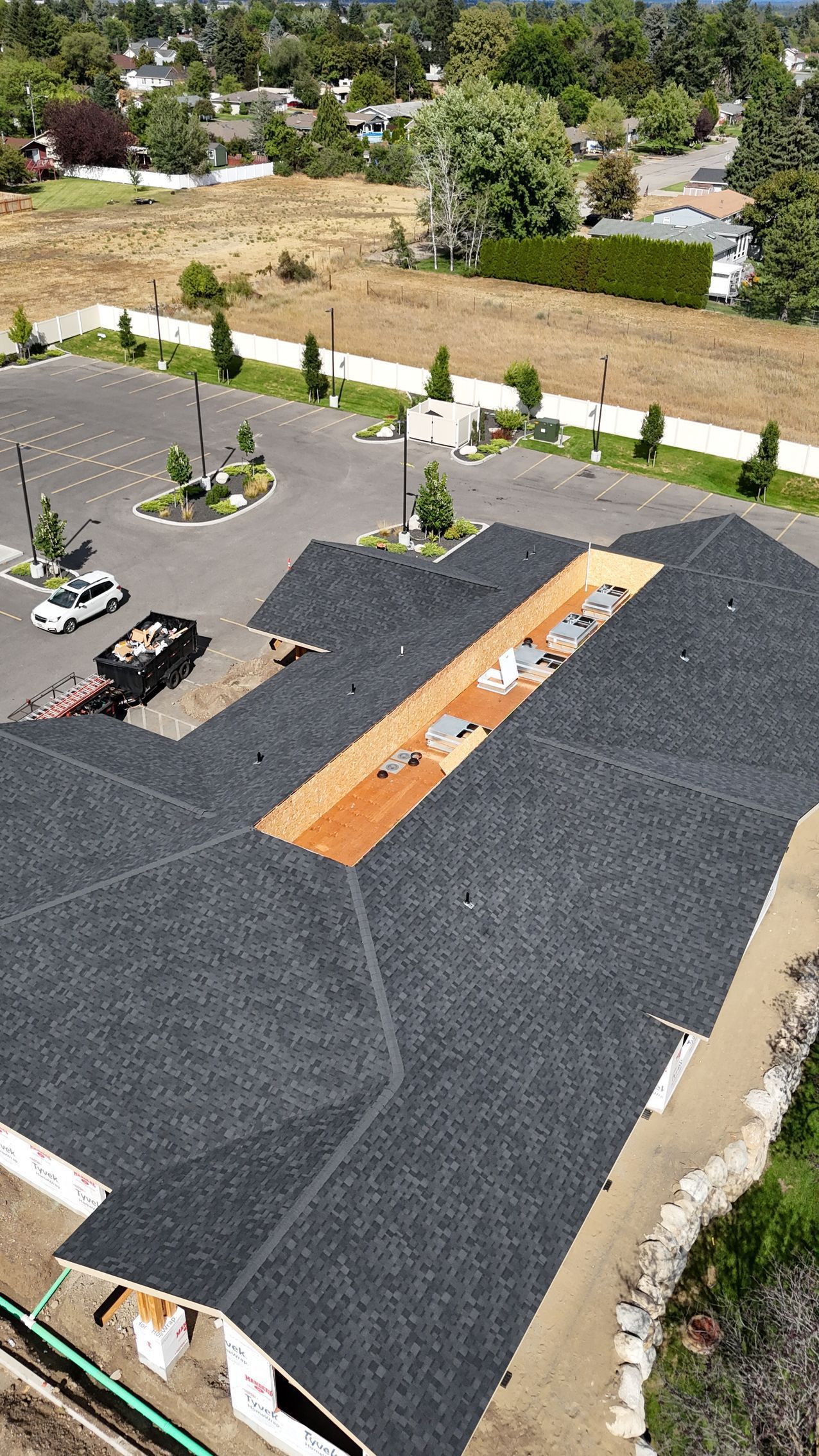 Aerial view of a building rooftop with dark gravel, a tan walkway, and parked cars beside a landscaped lot