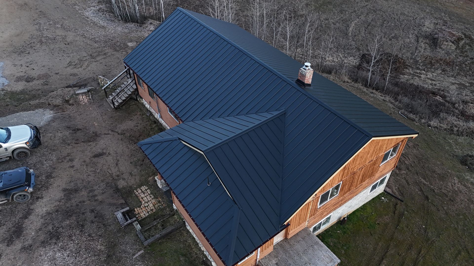 Aerial view of a house with a dark roof and orange brick walls beside a dirt driveway.