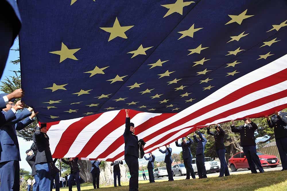 People holding a large American flag with gold stars and red-and-white stripes outdoors at a ceremony