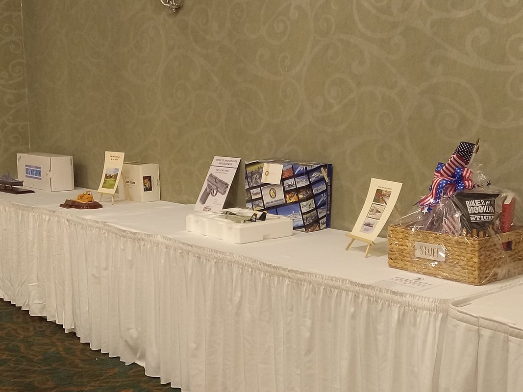 White-covered banquet table with raffle baskets, prizes, and signs arranged along a wall.