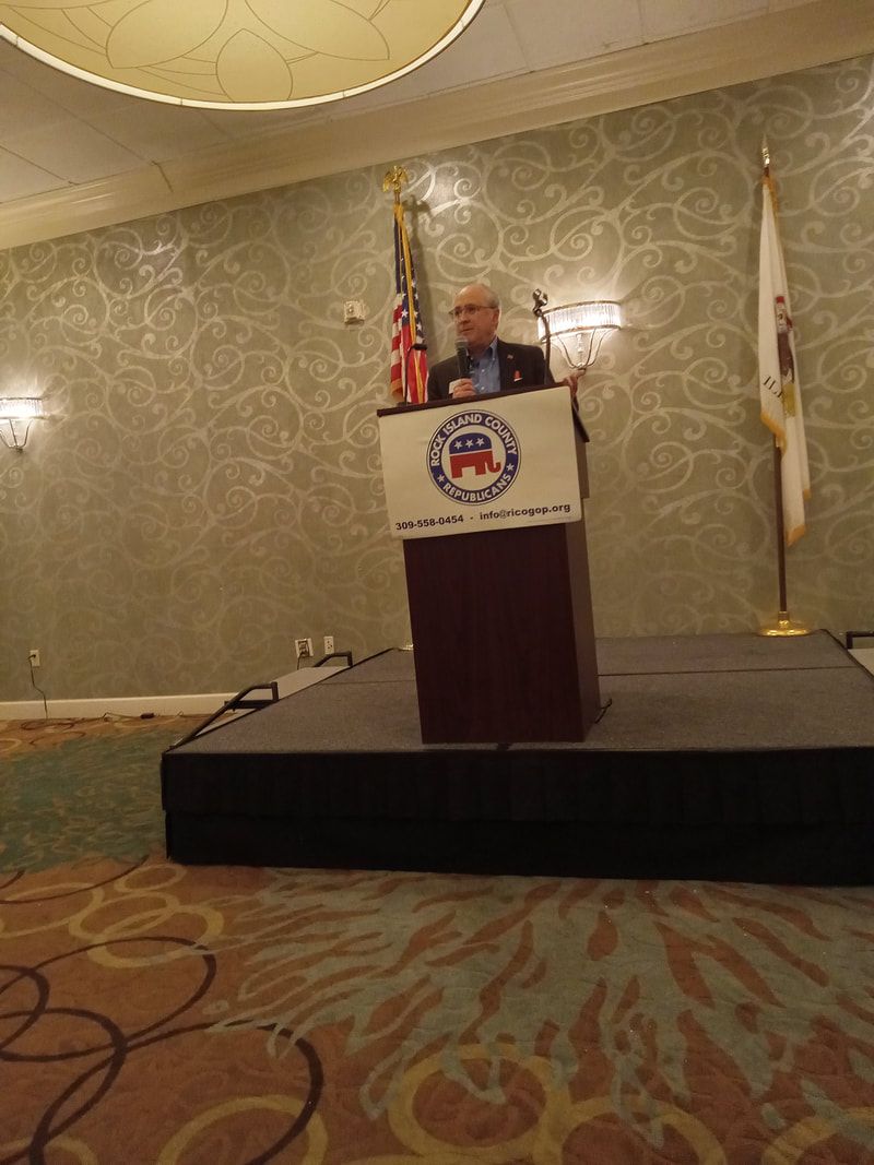 Man speaking at a podium on a stage in a banquet hall with flags behind him