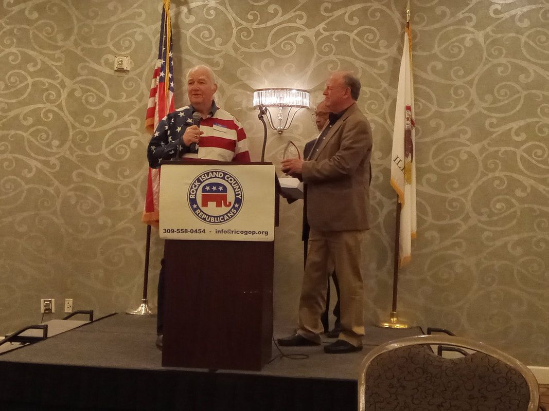 Two men at a podium with a patriotic emblem, speaking at an indoor event with U.S. and state flags.