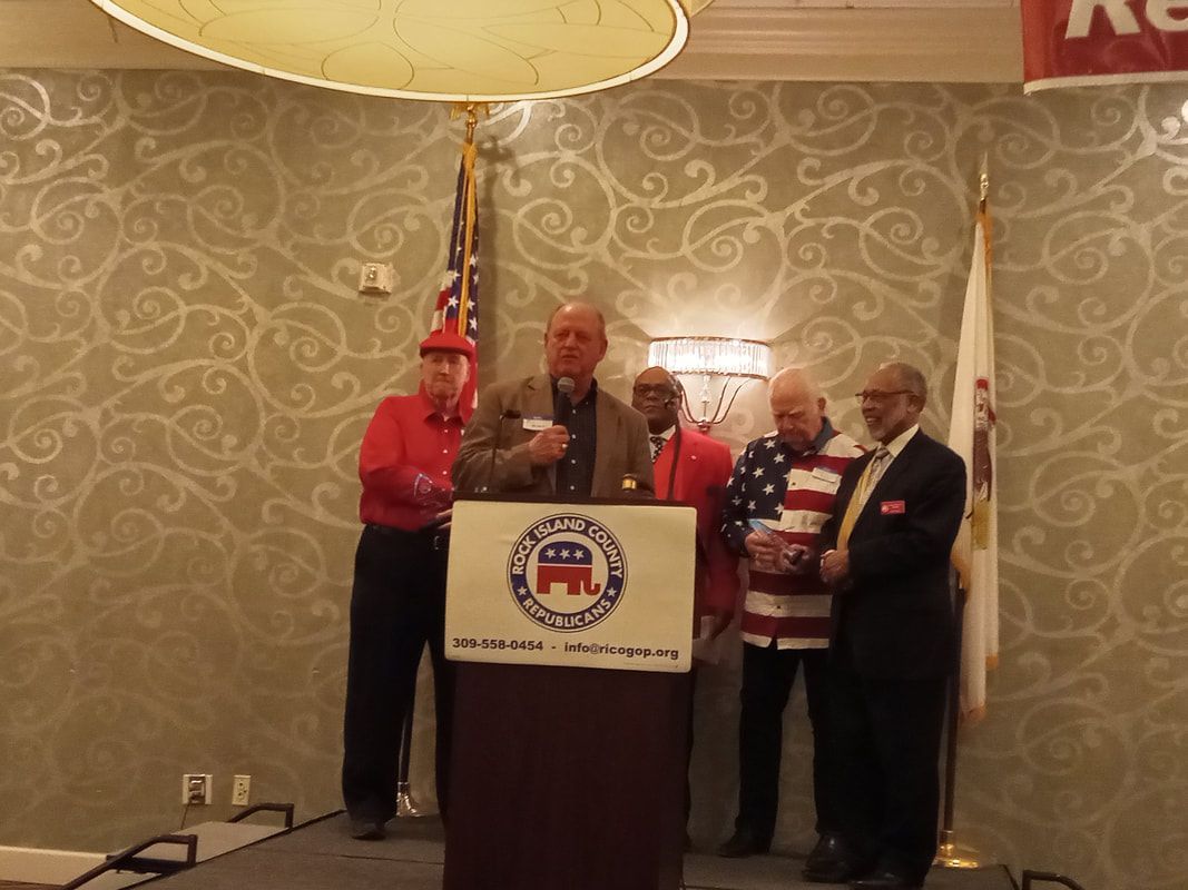 Man speaking at podium with four people standing beside him at a banquet-style event.
