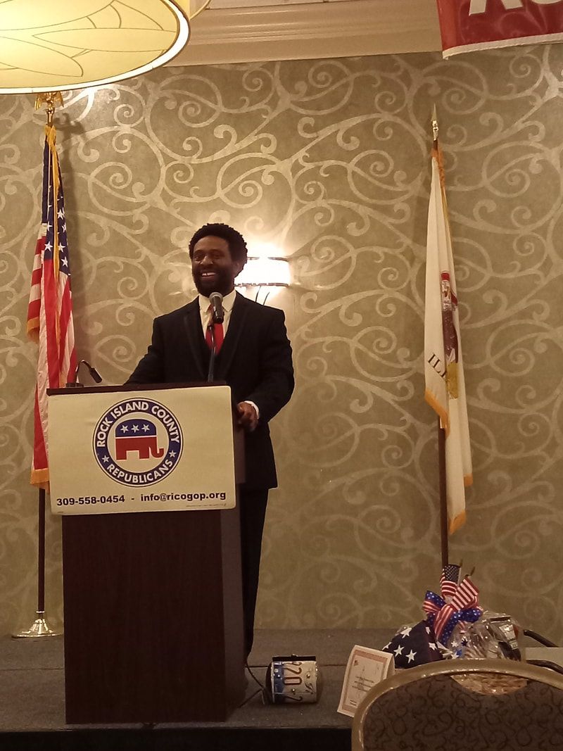 Man speaking at a podium in a decorated hall with U.S. and state flags behind him