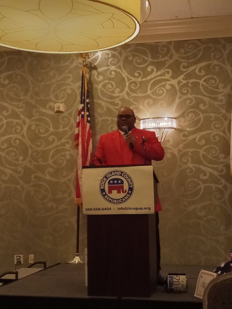 Man in red jacket speaking at a podium with an emblem, beside an American flag in a banquet room