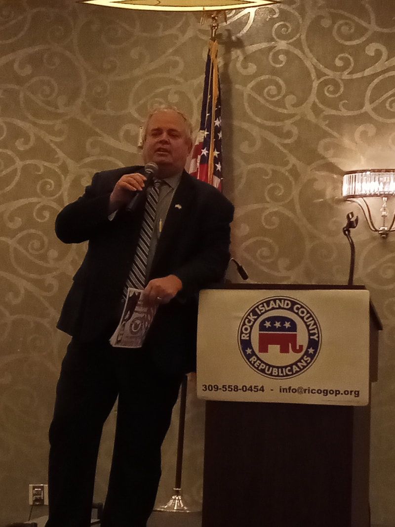 Man speaking at a podium beside an American flag in a hotel ballroom.