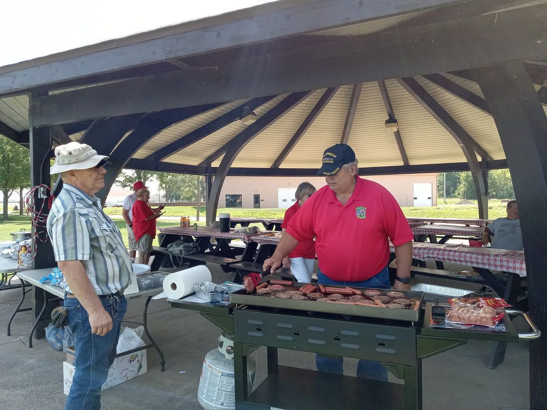 Two men tending a barbecue at a park picnic shelter, with tables and grills under a roof.