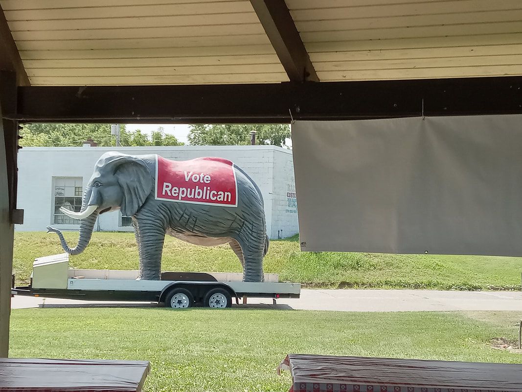 Elephant statue on a trailer, covered with a red Republic sign, under a pavilion with grass beyond.