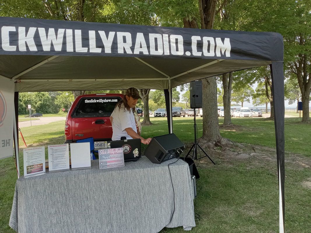 Person at a CKWILLYRADIO.com booth under a canopy, with boxes, speaker, and a red vehicle behind.