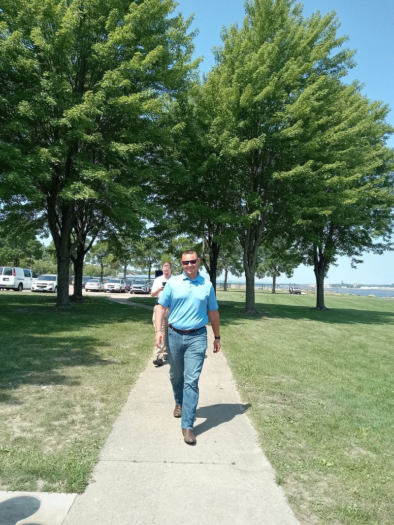 Person walking on a sidewalk in a park with trees and parked cars in the background