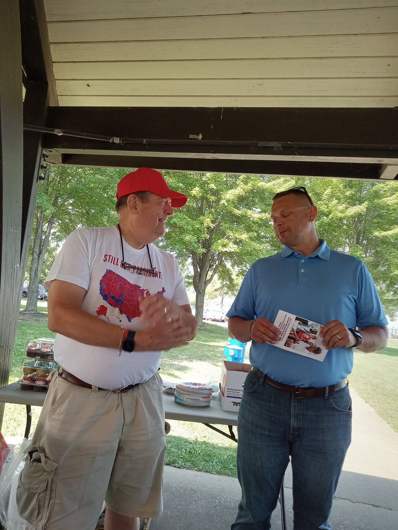 Two men standing under a pavilion, talking and holding papers, with trees and parked cars behind them.
