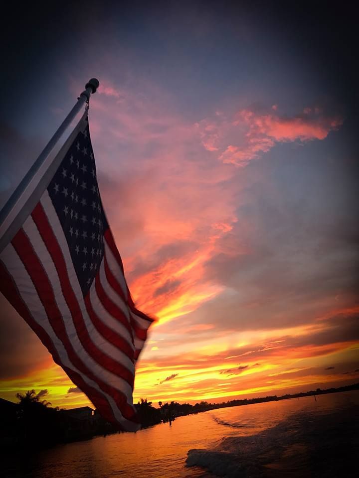 American flag waving at sunset over water, with a vivid orange and pink sky