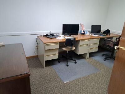 Office desk area with computer monitors, chairs, and filing drawers against a plain wall.