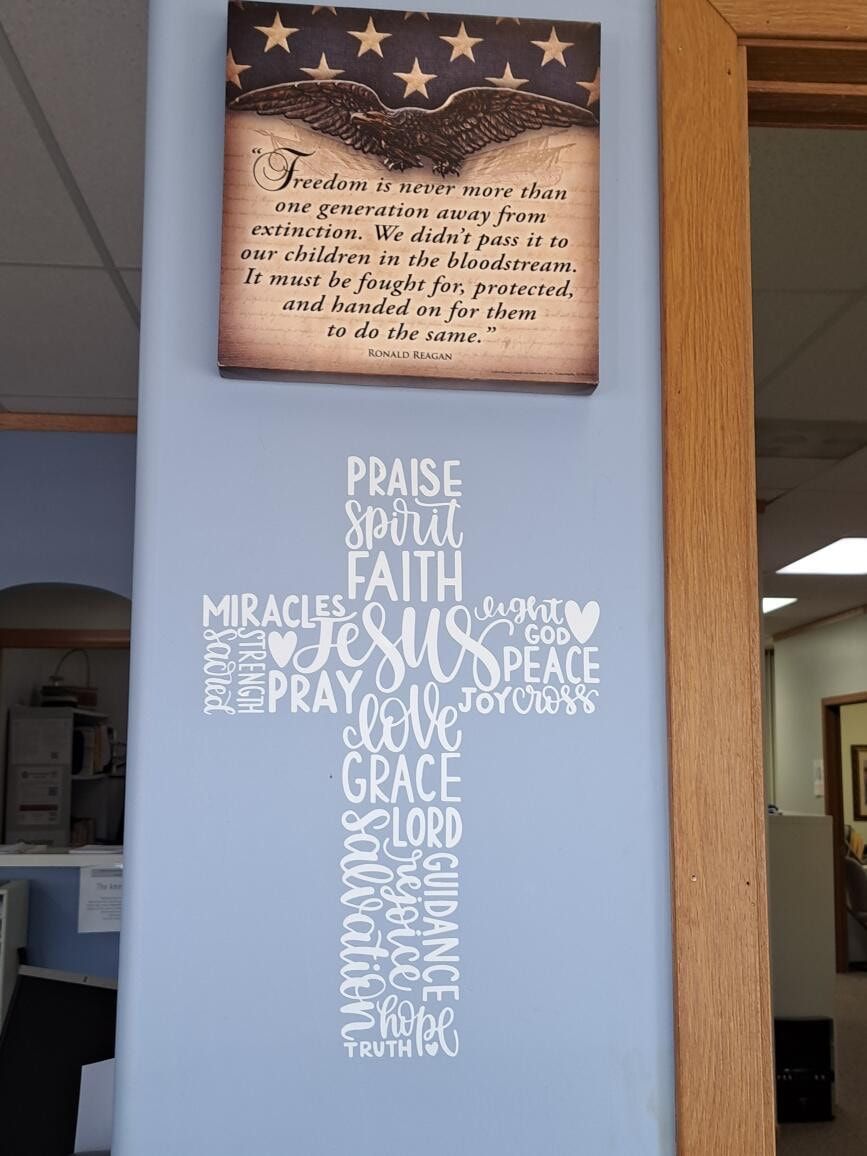 Framed patriotic sign above white wall decal cross with faith words in a home hallway