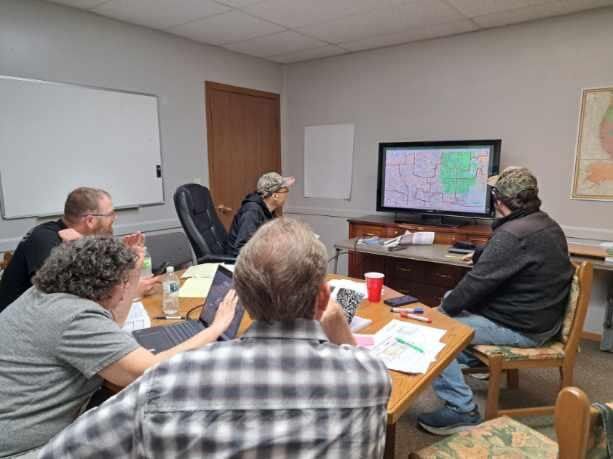 People in a meeting room watching a large screen with maps during a presentation.