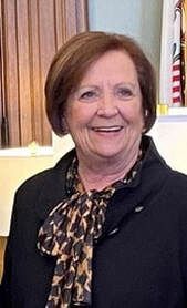Smiling woman in a black jacket and patterned scarf standing indoors, with curtains and a flag behind her