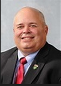 Portrait of a smiling bald man in a dark suit, light blue shirt, and red tie against a gray background