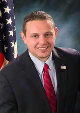 Official portrait of a man in a dark suit and red tie, standing before a U.S. flag backdrop
