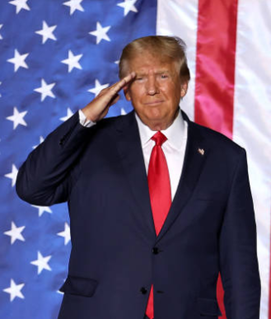 Man in a dark suit and red tie saluting before a U.S. flag backdrop