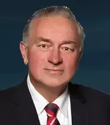 Formal headshot of a man in a dark suit and red striped tie against a blue background