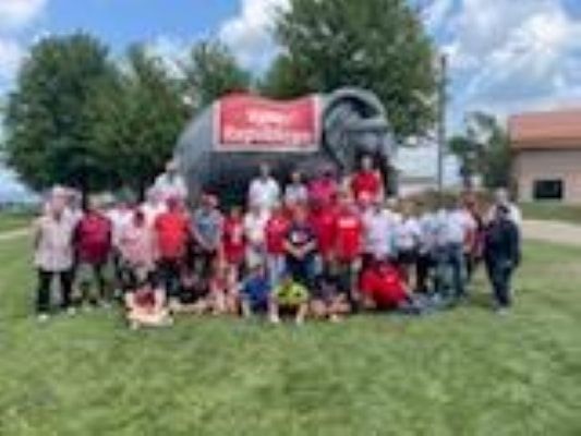 Group posing on a grassy field in front of a red inflatable event arch and tents outdoors.