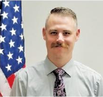 Man in dress shirt and patterned tie standing beside a U.S. flag against a plain background