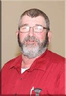 Man in red shirt with glasses and gray beard, standing against a beige wall.