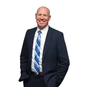 Smiling bald man in a navy suit with blue striped tie, standing against a white background