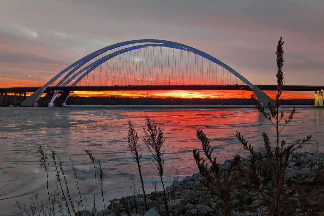 Blue arch bridge at sunset over a river, with orange sky and silhouetted reeds in the foreground