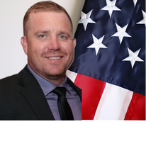 Man in suit beside a U.S. flag, smiling at the camera.