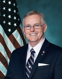 Smiling man in suit and tie standing before a U.S. flag backdrop