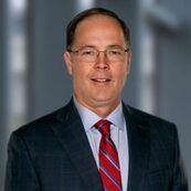 Head-and-shoulders portrait of a man in a dark suit, red striped tie, and glasses on a gray background