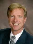 Smiling man in a dark suit, striped tie, and white shirt against a studio backdrop