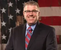 Man in suit and striped tie standing before a U.S. flag, smiling