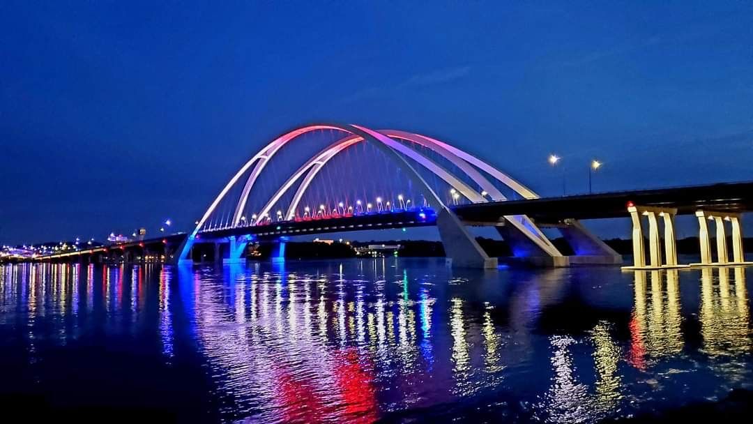 Illuminated arched bridge glowing pink and blue over calm water at night, with reflections and city lights