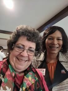 Two smiling people posing indoors, one in a colorful scarf, one in a red jacket with a name tag.