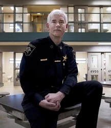 Uniformed officer sitting on a bench in a corridor, with glass windows and doors behind him.