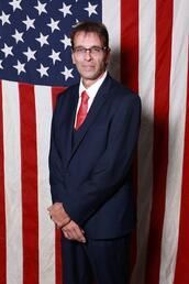 Man in dark suit and red tie standing before a large American flag backdrop