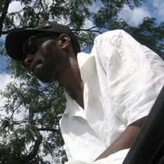 Man in a white shirt and black cap, viewed from below against a leafy sky.