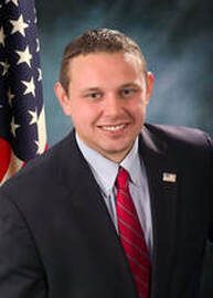 Man in dark suit and red tie posing beside an American flag against a studio backdrop