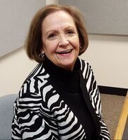 Smiling woman in a black turtleneck and zebra-striped cardigan seated indoors.