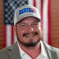 Smiling man in a gray jacket and baseball cap in front of an American flag backdrop