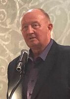 Man speaking at a microphone in a dark blazer and purple shirt, standing before a patterned wall.