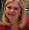 Smiling blonde woman in a purple top, seated indoors against a neutral background.