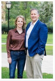 Two people standing outdoors, one in a brown blouse and jeans, the other in a blue blazer and khakis.
